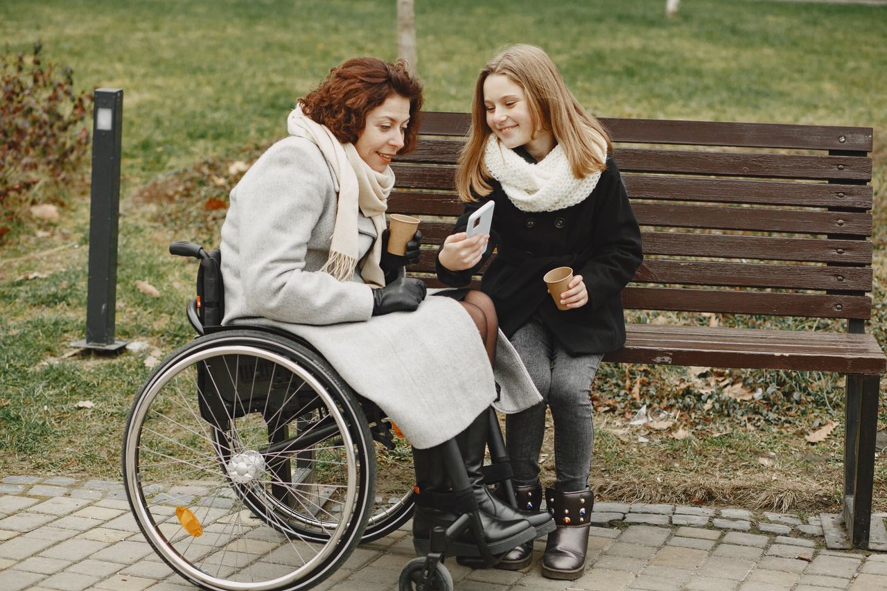 A woman in a wheelchair and her daughter sharing a joyful moment outdoors with a mobile phone.
