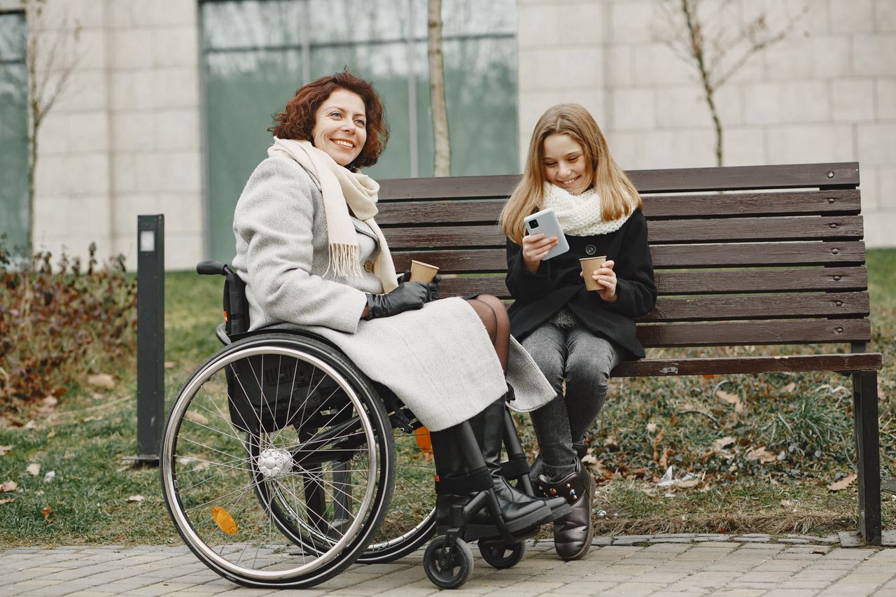 A joyful moment between mother in wheelchair and daughter sharing coffee on a park bench.