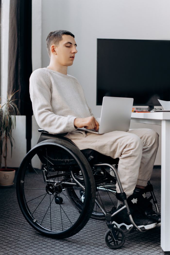Young man in wheelchair using laptop indoors, illustrating inclusivity and technology use.