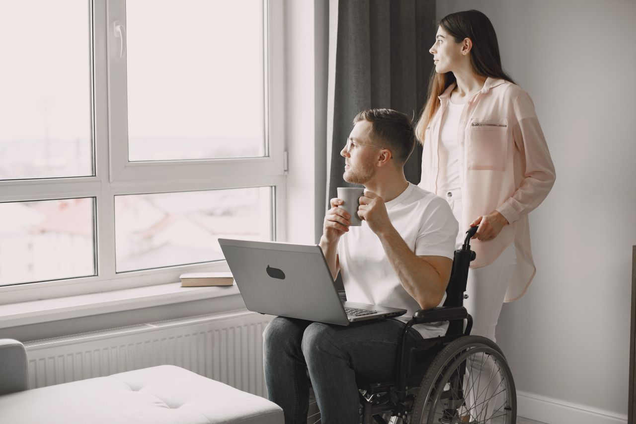Man in wheelchair with laptop and coffee, gazing out window with a woman beside him.