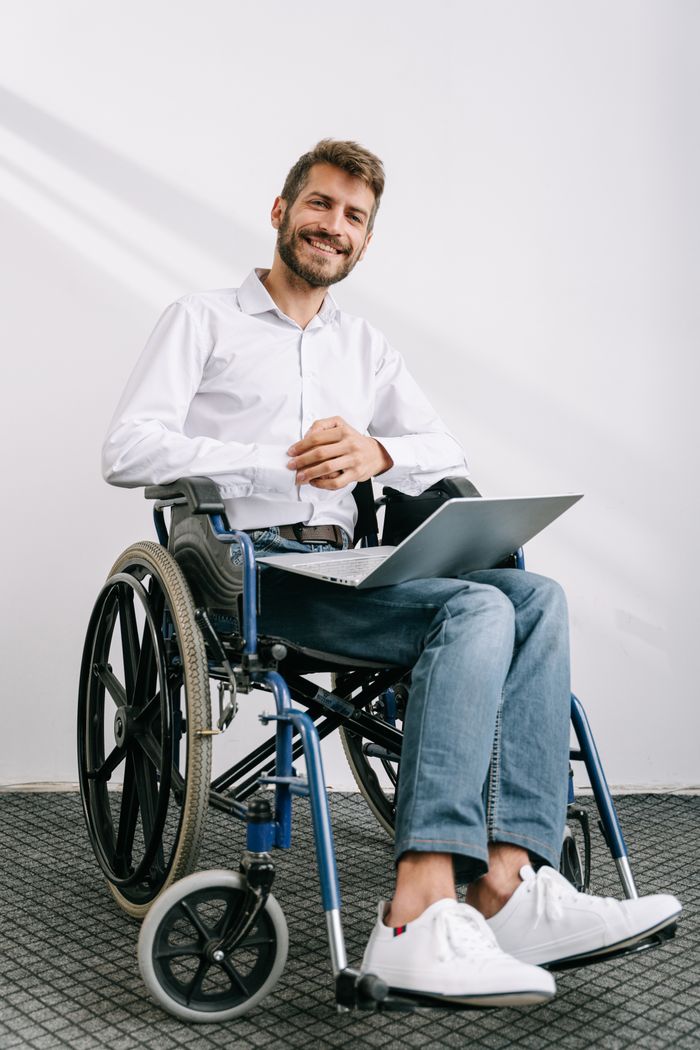 Confident bearded man in wheelchair using laptop while smiling in a bright office setting.
