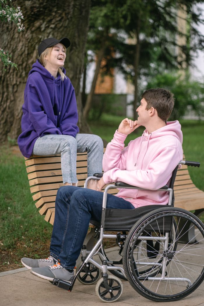 A young couple enjoying a cheerful conversation outdoors, dressed casually with a wheelchair in view.